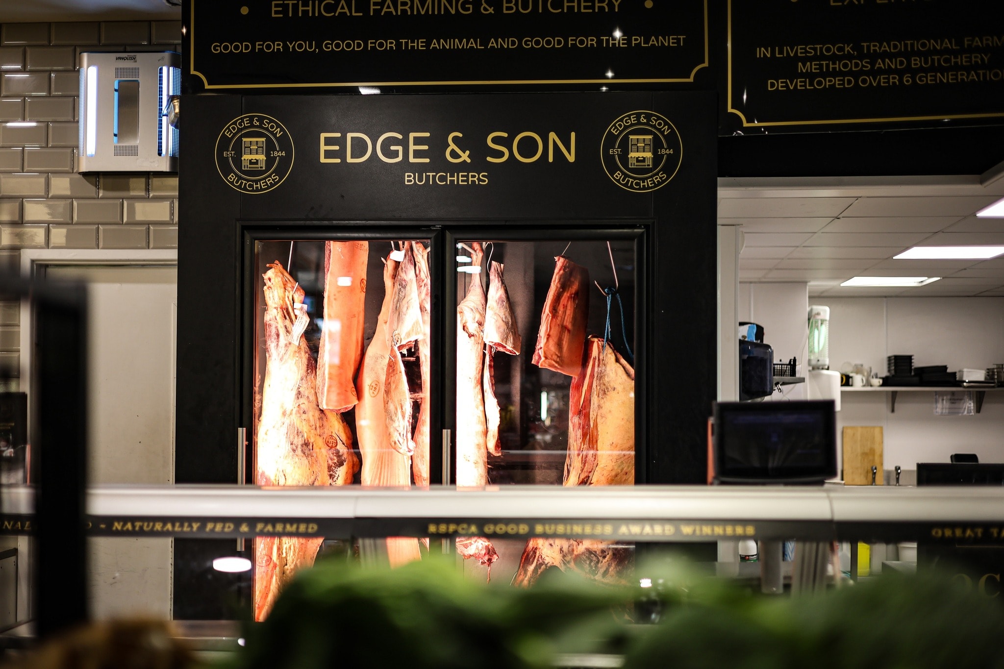 Joints of meat hanging in the lit display fridge behind the Edge & Son counter