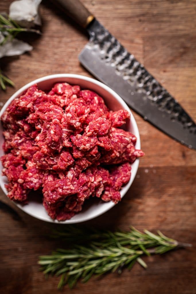 A white bowl of fresh minced beef on a wooden board with a knife and rosemary